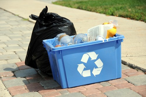 On a paved sidewalk in an outdoor urban setting, a blue plastic recycling bin with a white recycling symbol on its side is filled with empty plastic bottles, some with labels and caps, and a white container with a yellow cap. Next to the bin, a large black garbage bag, tightly sealed, is leaning against it. The background includes a grassy area and a curb, indicating a residential or commercial environment in Newham. The scene illustrates typical rubbish collection involving recyclable materials and general waste, consistent with waste removal services offered by companies like Commercial Waste Removal Newham, serving local postcode areas.