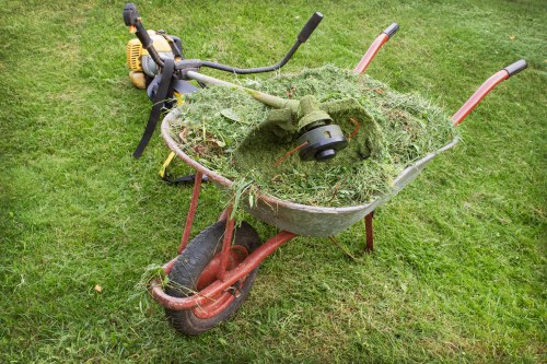 A red and black wheelbarrow filled with freshly cut grass and yard waste, positioned on a green lawn surface. The wheelbarrow contains a mix of grass clippings, small plant debris, and soil, with the handles extending towards the background. In the background, a petrol-powered grass trimmer with a black handle and yellow engine housing is resting on the grass. The scene appears to be outdoors, likely in a residential garden or yard within Newham, suggesting the need for rubbish removal services for garden waste by Commercial Waste Removal Newham. The environment is well-lit, emphasizing the freshly cut vegetation and outdoor setting.