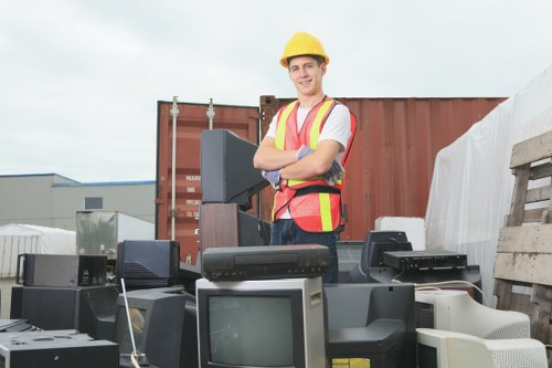 A man wearing a yellow safety helmet and a high-visibility vest with reflective stripes is standing outdoors in an industrial or warehouse yard setting, surrounded by various discarded electronic devices such as old computer monitors, printers, and other outdated office equipment. The background features large shipping containers, with one prominently visible in rust-brown color, and a partly cloudy sky overhead. The man is smiling and has his arms crossed, appearing confident and professional. The electronic waste is piled on a paved surface and appears to be abandoned or prepared for removal. This scene illustrates a rubbish removal service in action, focusing on electronic waste collection, and the environment hints at a location in Newham or nearby areas, emphasizing the services provided by Commercial Waste Removal Newham.