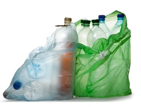 A collection of various transparent plastic bottles positioned on a flat surface, including a large crumpled white plastic bag, with bottles of different shapes and sizes, some with caps and others without. The bottles appear to be made from PET material, featuring smooth, glossy surfaces with visible ridges and slightly varying shades of clear, green, and blue. The background is plain white, emphasizing the items which are situated in a somewhat clustered arrangement, with the largest bag slightly leaning to the left and the bottles stacked or placed upright within the bag. The arrangement suggests a typical scene of plastic bottle recycling or rubbish for collection by a waste removal service like Commercial Waste Removal Newham, highlighting waste segregation along with environmental responsibility in a residential or commercial setting in London or the surrounding area.
