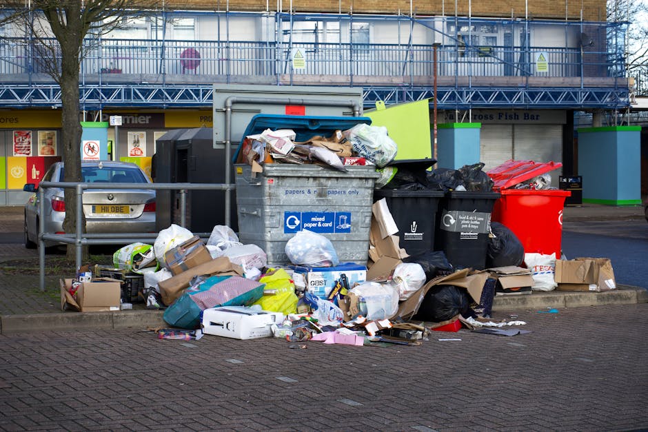 A close-up view of three large black plastic rubbish bags resting on a curbside pavement in an outdoor urban environment during daytime. The bags are tightly packed and appear to contain household was