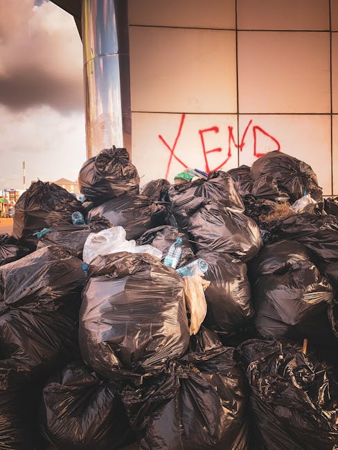 Close-up view of a tightly tied black plastic rubbish bag with a glossy surface, resting against other similar bags in a pile. The bags are placed outdoors on a paved or concrete surface, with a plain